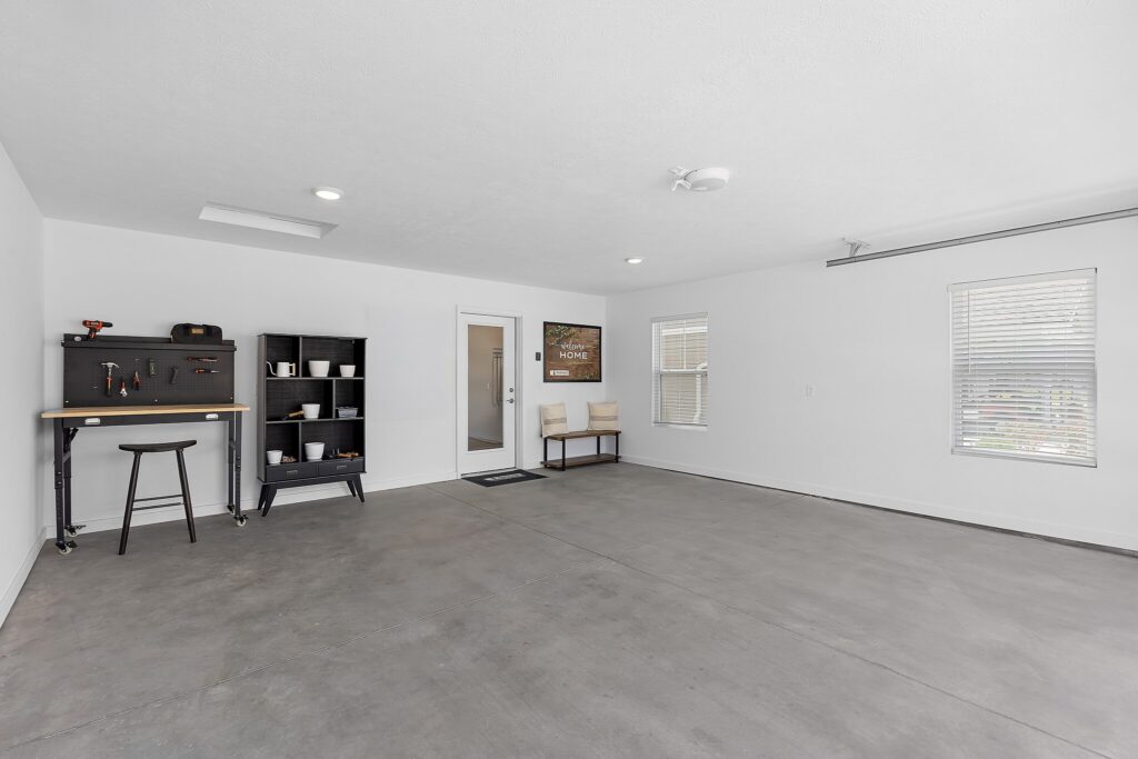 Spacious garage with gray floor, white walls, workbench, shelves, bench, and two windows letting in natural light.