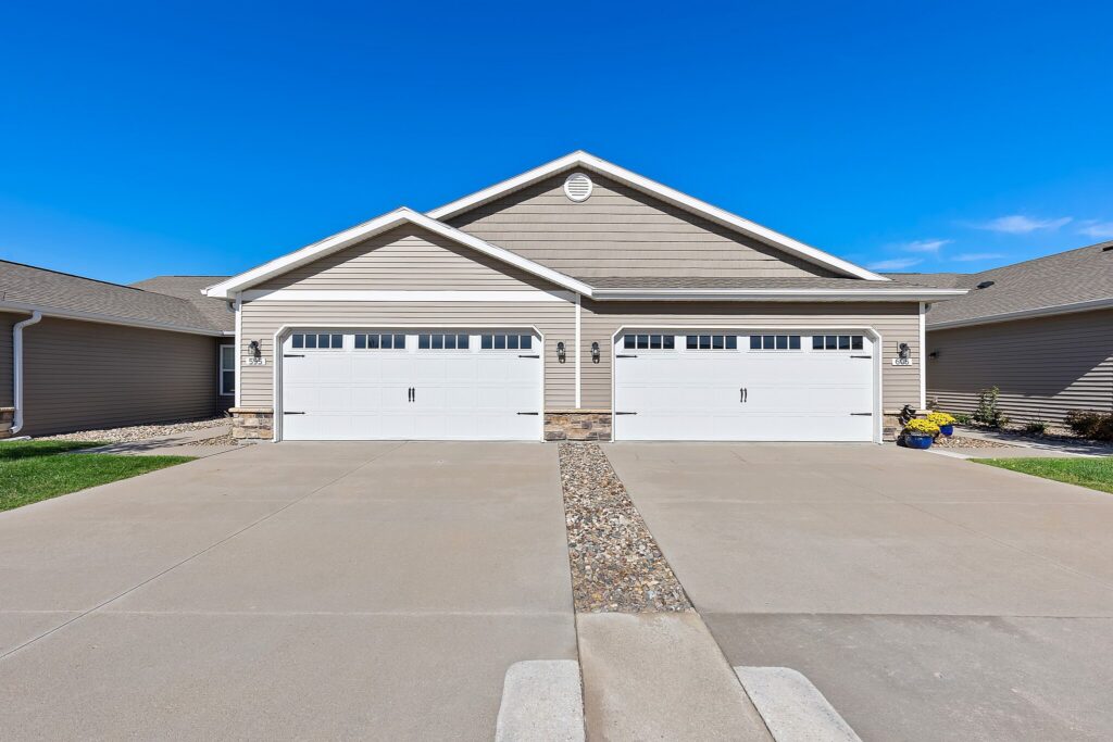 A duplex with two white garage doors, tan siding, and a shared concrete driveway under a clear blue sky.