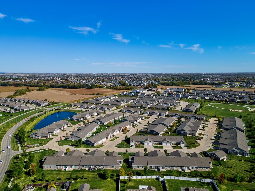Aerial view of a suburban neighborhood with rows of houses, green lawns, a small pond, and open fields under a clear blue sky.