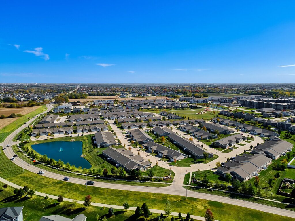 Aerial view of a suburban neighborhood with rows of houses, a pond with a fountain, green lawns, and roads under a clear blue sky.