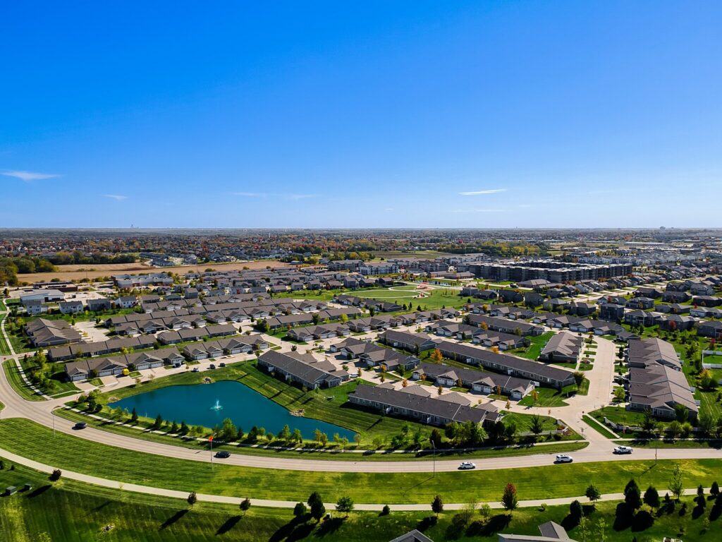 Aerial view of a suburban neighborhood with houses, roads, a pond with a fountain, and green lawns under a clear blue sky.