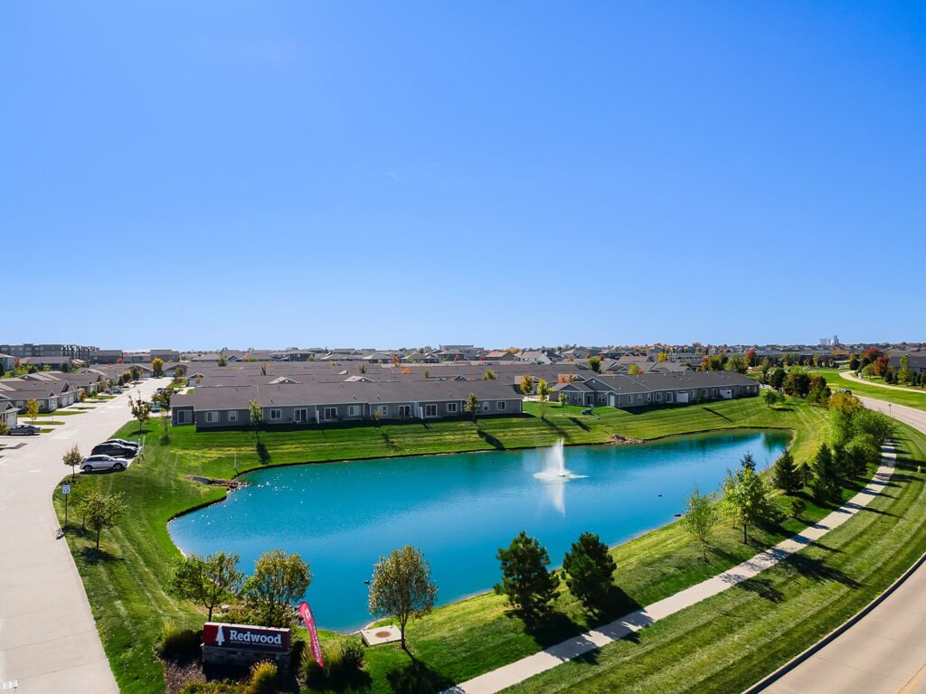 A residential community surrounds a pond with a fountain. Well-maintained grass, trees, and a visible sign reading “Redwood” are present along the paved roadway.