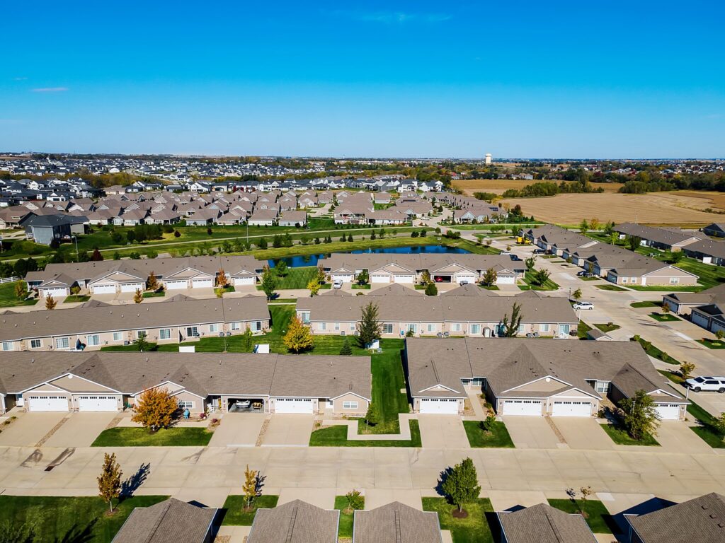 Aerial view of a suburban neighborhood with rows of houses, green lawns, driveways, and a small pond in the center, surrounded by open fields under a clear blue sky.