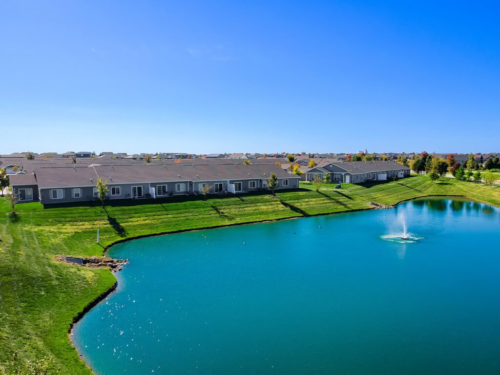 Row of single-story houses with gray siding overlooks a pond with a central fountain, surrounded by green grass and a clear blue sky.
