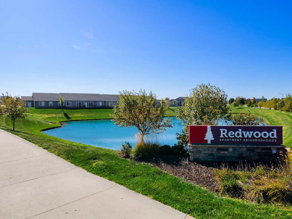 A sidewalk runs beside a pond, landscaped grass, and trees, with a “Redwood Apartment Neighborhoods” sign in the foreground and single-story buildings in the background.