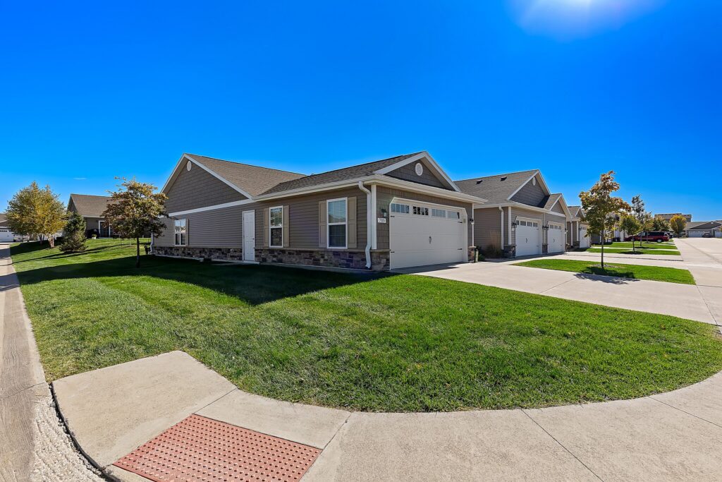 Single-story suburban house with attached garage, well-maintained lawn, and clear blue sky; similar homes line the residential street.