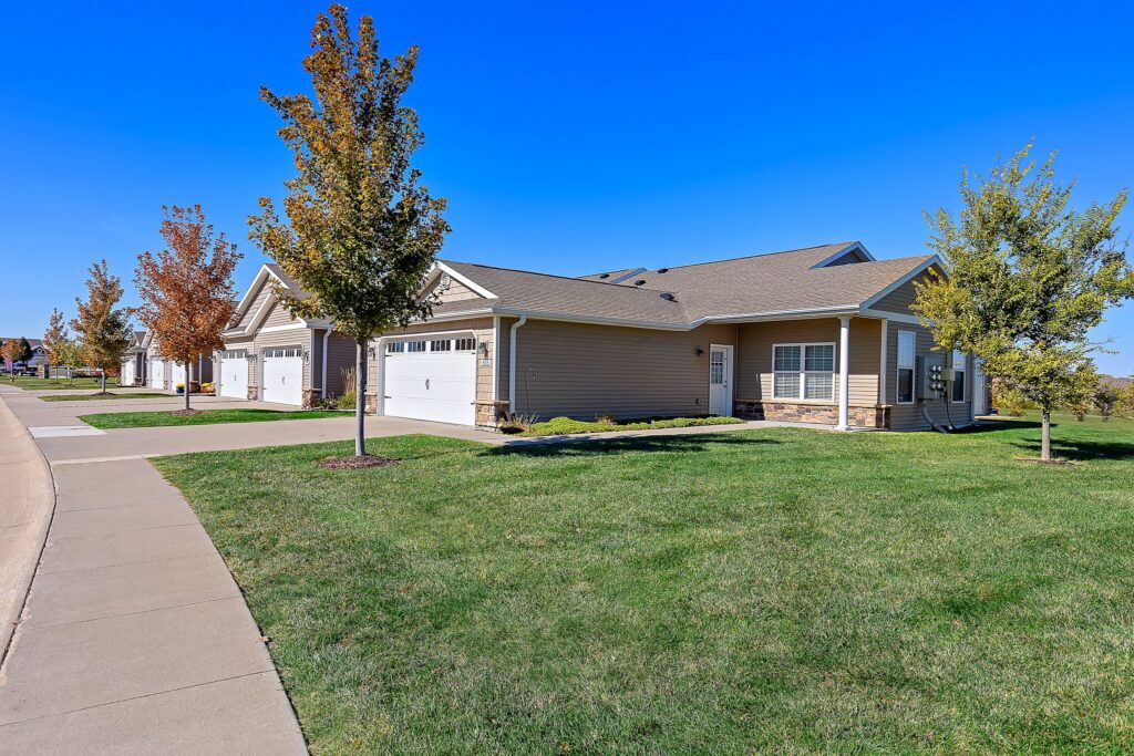 Single-story suburban house with attached garage, beige siding, manicured lawn, young trees, and a clear blue sky. Concrete driveway and sidewalk visible in the foreground.