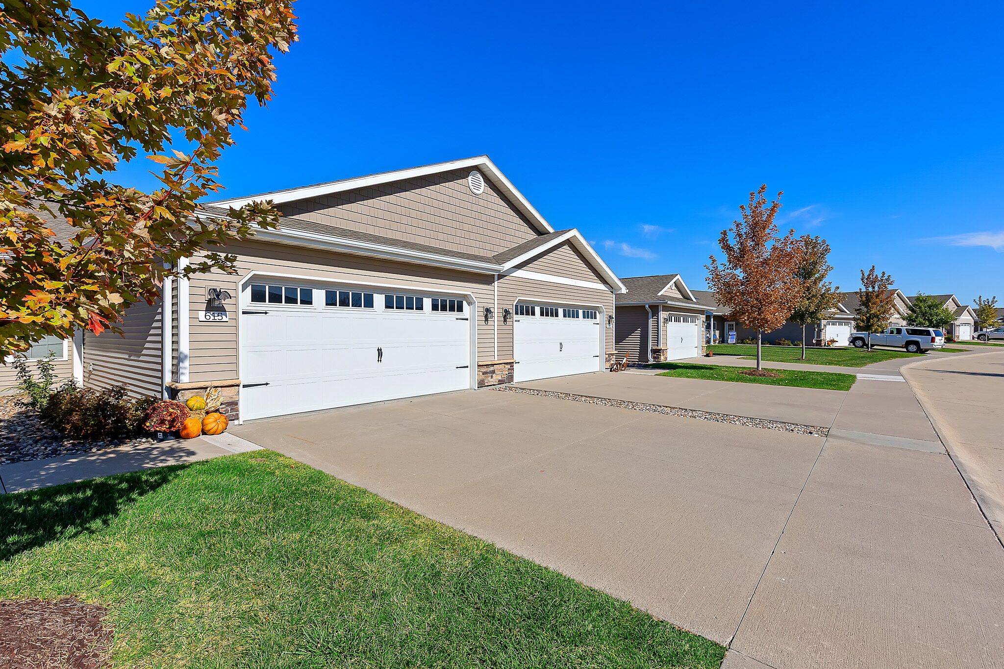 Row of beige suburban houses with attached white garages, concrete driveways, green lawns, and a few autumn trees on a clear, sunny day.