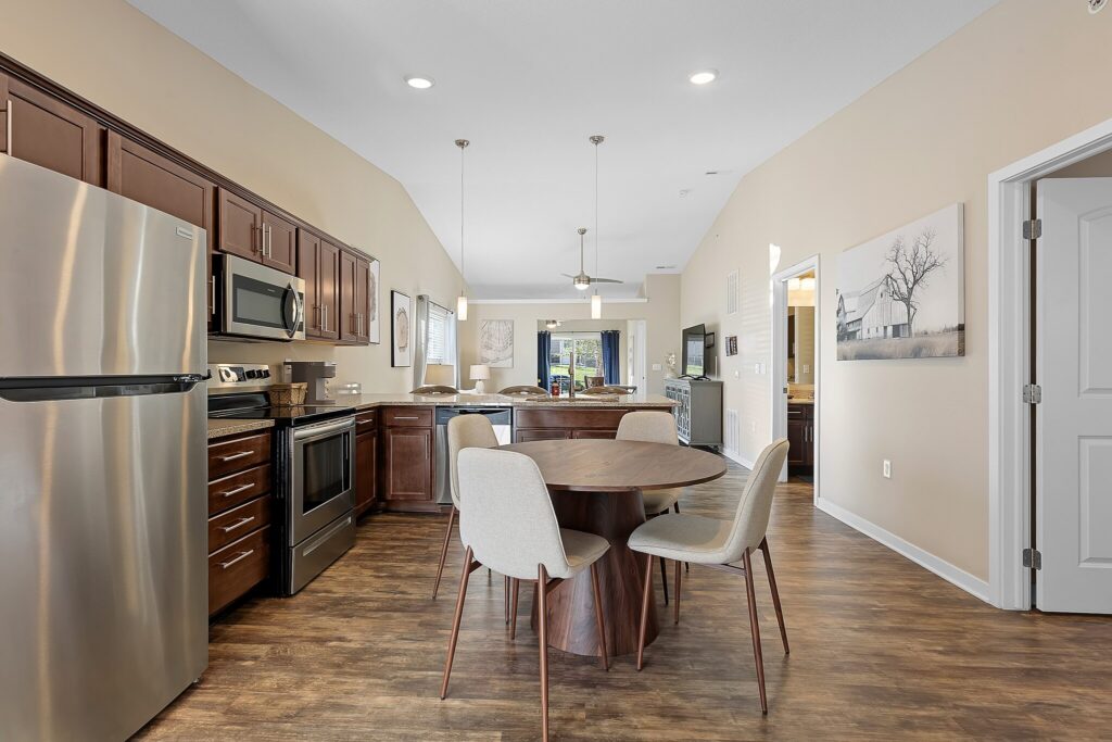 Modern kitchen and dining area with stainless steel appliances, wooden cabinets, round dining table with four chairs, and wood flooring. Living area visible in the background.