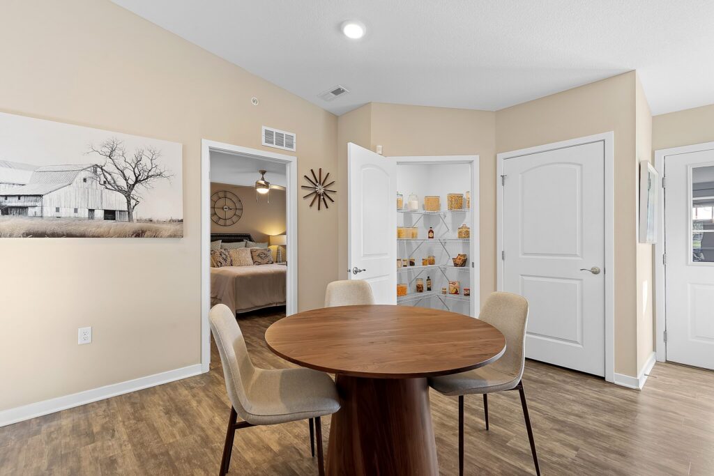A round wooden table with three chairs sits in a beige room. The image shows doors to a bedroom, a pantry with shelves, and another room. Wall art and a clock decorate the walls.