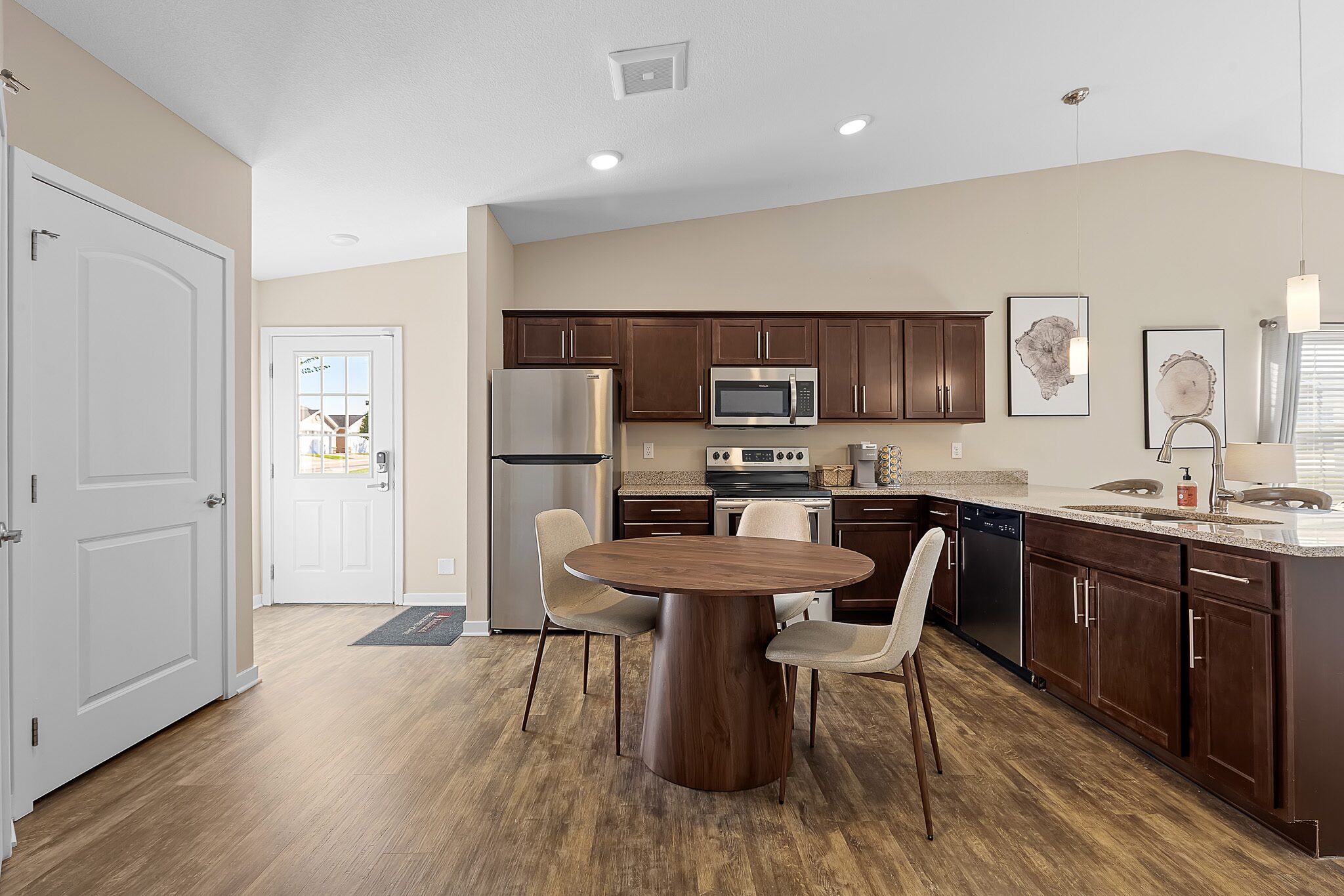 Modern kitchen with dark wood cabinets, stainless steel appliances, a round dining table with three chairs, and wood-look flooring. Natural light enters through a door and window.