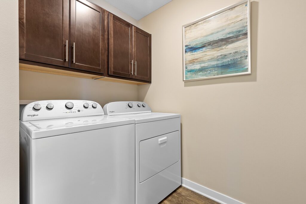 A laundry room with a white washer and dryer, brown cabinets above, beige walls, wood floor, and a framed abstract painting on the wall.