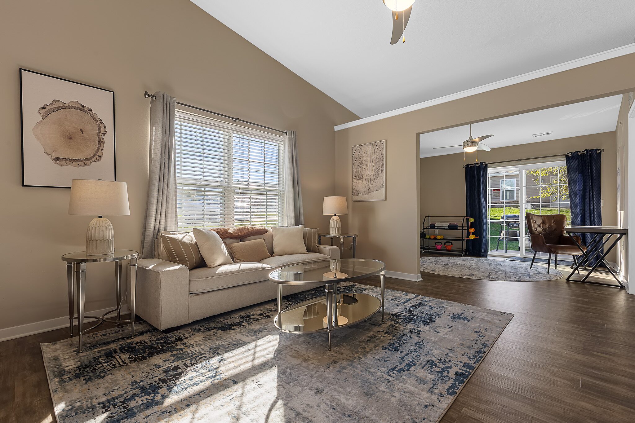 Living room with beige walls, a cream sofa, glass coffee table, two lamps, and large window; adjoining room with desk, dark curtains, and child’s play area, all with wood flooring.