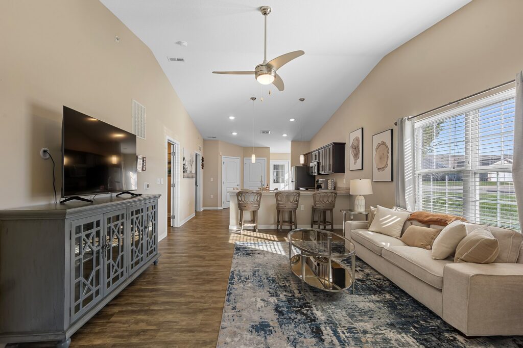 Open-concept living room and kitchen with beige walls, large window, modern furniture, TV, and wooden flooring.