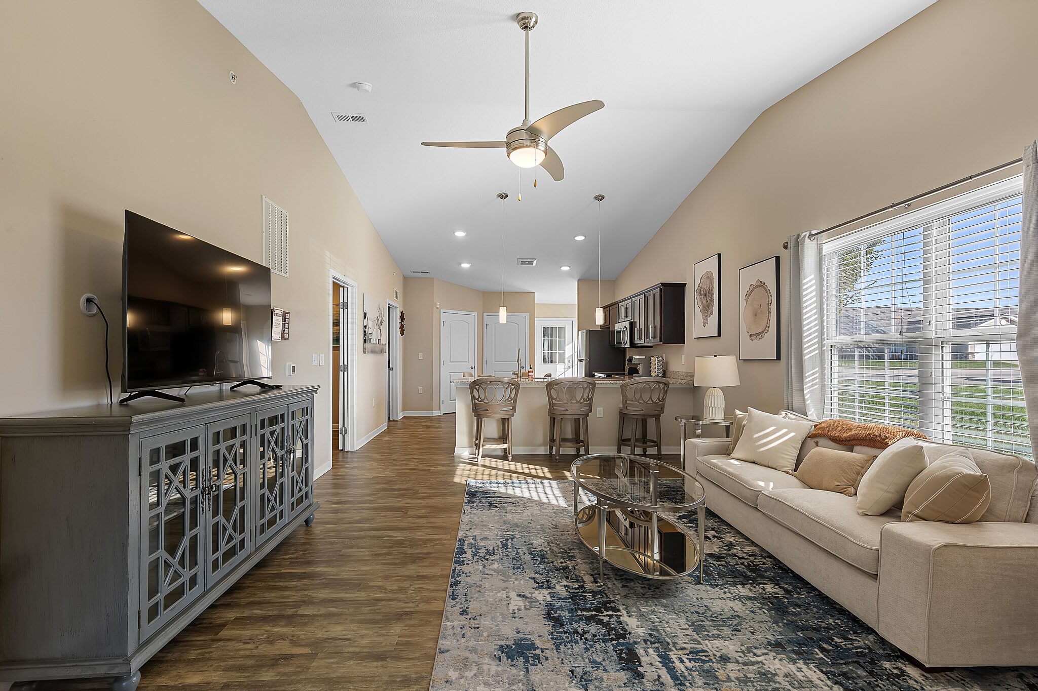 Open-concept living room and kitchen with beige walls, large window, modern furniture, TV, and wooden flooring.