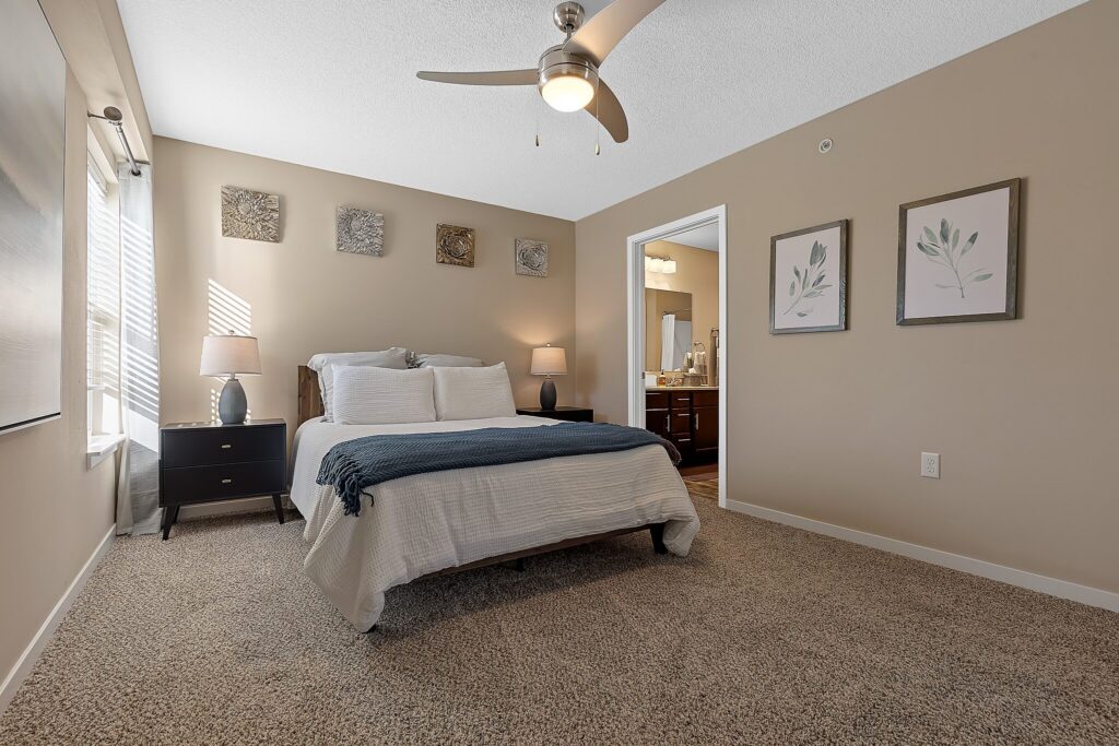 A neatly made bed with white and gray bedding sits in a carpeted bedroom with beige walls, nightstands, lamps, wall art, and a ceiling fan. An open door leads to a bathroom.