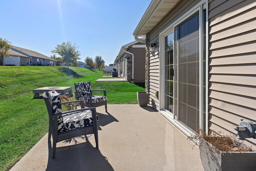 Two patterned chairs and a small table sit on a concrete patio outside a beige house, with a grassy yard and other houses visible in the background under a clear sky.