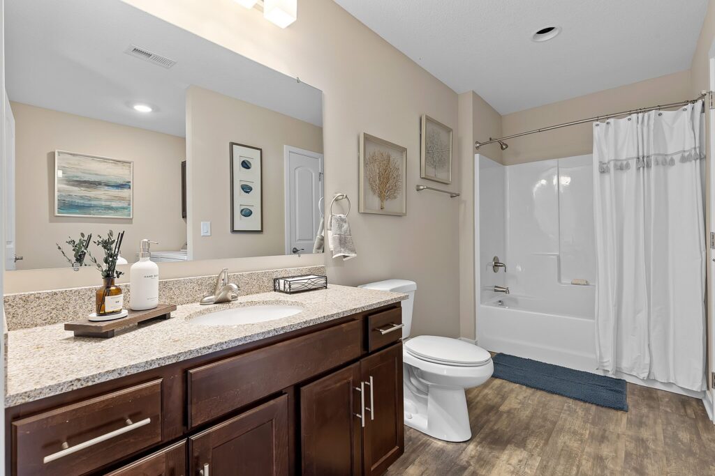 Modern bathroom with a granite countertop, dark wood cabinets, white sink, toilet, wall art, and a shower-tub combo with a white curtain on a wooden floor.