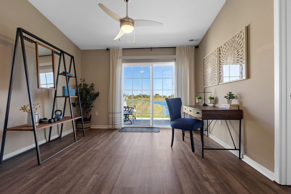 Home office with a wooden desk, blue chair, open shelving unit, plant, ceiling fan, and large sliding glass doors letting in natural light.