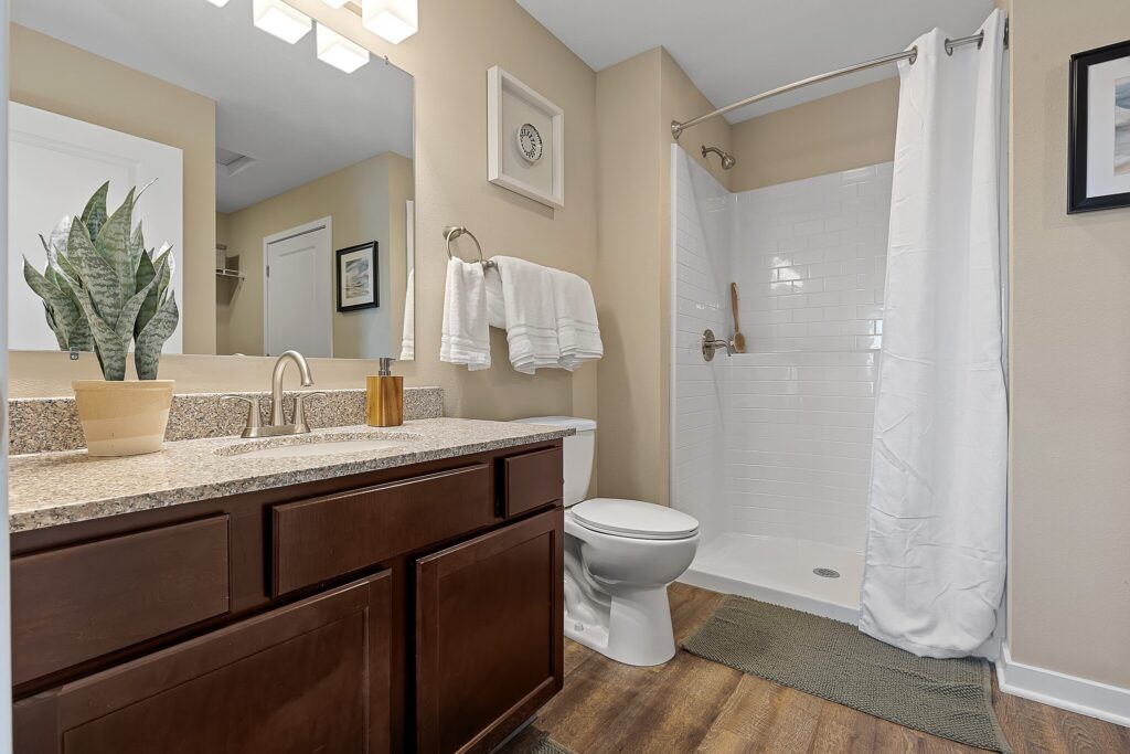 Modern bathroom with granite countertop, dark wood vanity, potted plant, white shower with curtain, toilet, and towels on a rack against beige walls.
