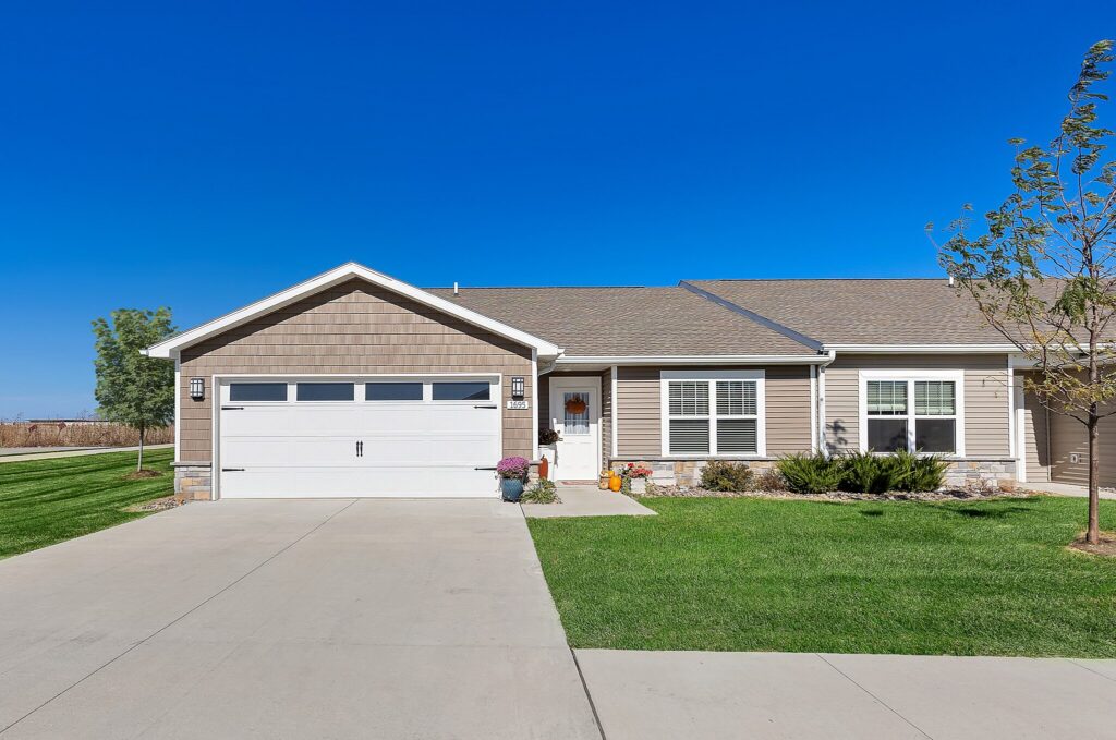 Single-story suburban house with a double garage, beige siding, manicured lawn, and a clear blue sky in the background.