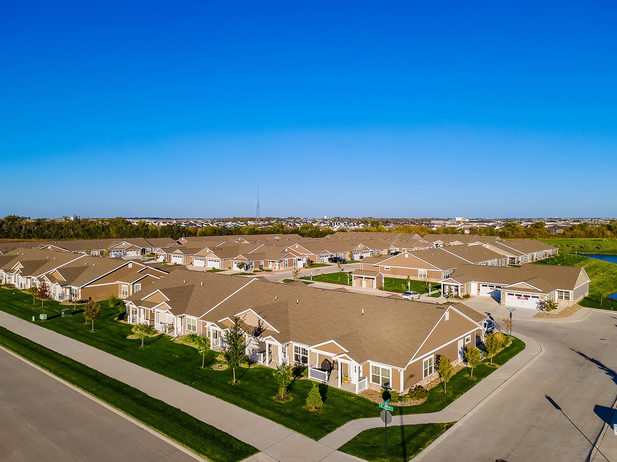 A suburban neighborhood with rows of single-story houses, paved streets, green lawns, and clear blue sky, seen from above.