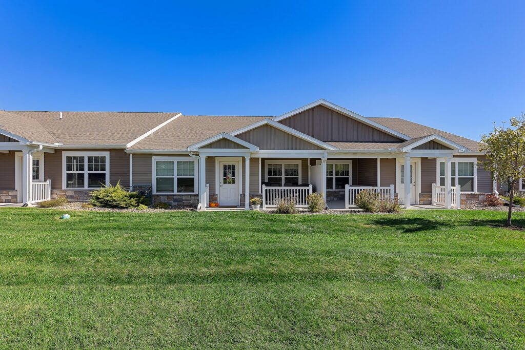 Single-story townhouse units with beige siding, white trim, covered porches, and a well-maintained green lawn under a clear blue sky.