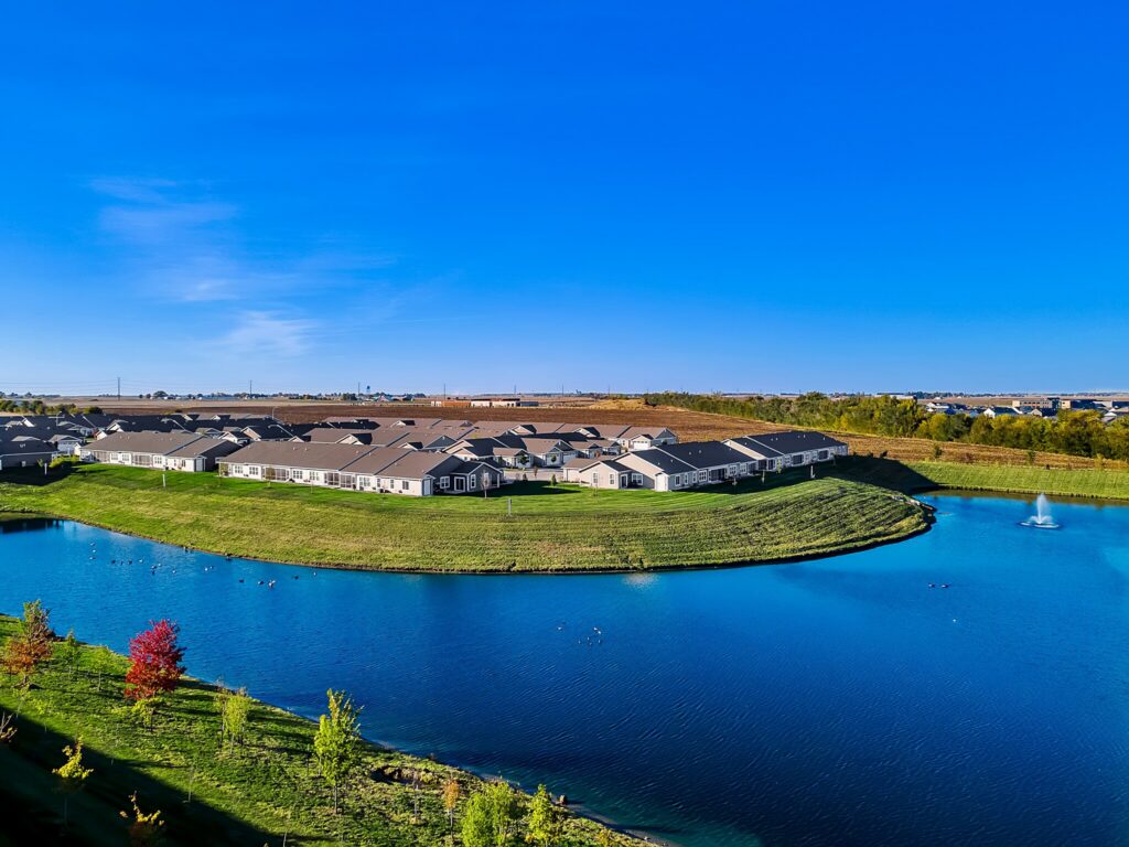 A cluster of houses sits on a grassy peninsula surrounded by a blue pond with a fountain, under a clear blue sky.