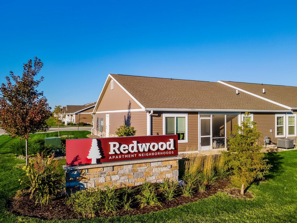 A red sign reads “Redwood Apartment Neighborhoods” in front of a single-story apartment building with a manicured lawn and clear blue sky.