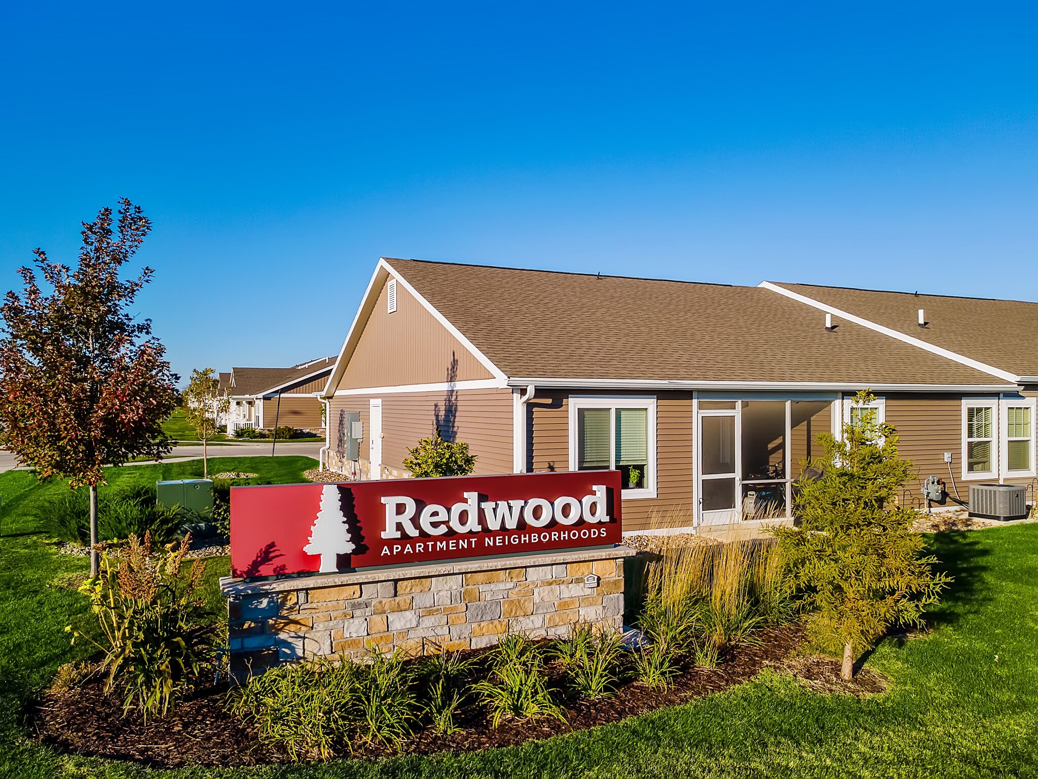 A red sign reads “Redwood Apartment Neighborhoods” in front of a single-story apartment building with a manicured lawn and clear blue sky.