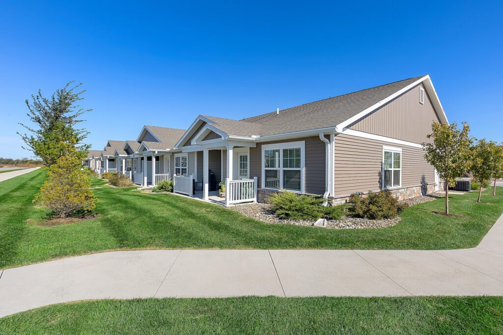 Single-story suburban houses with front porches line a sidewalk, surrounded by green lawns and young trees under a clear blue sky.