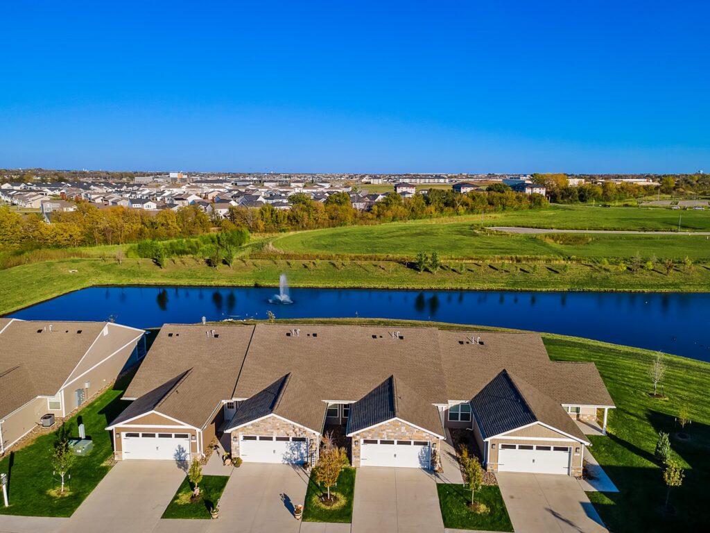 Aerial view of suburban houses with garages facing a pond, green lawns, and a fountain, with a larger neighborhood visible in the background under a clear blue sky.
