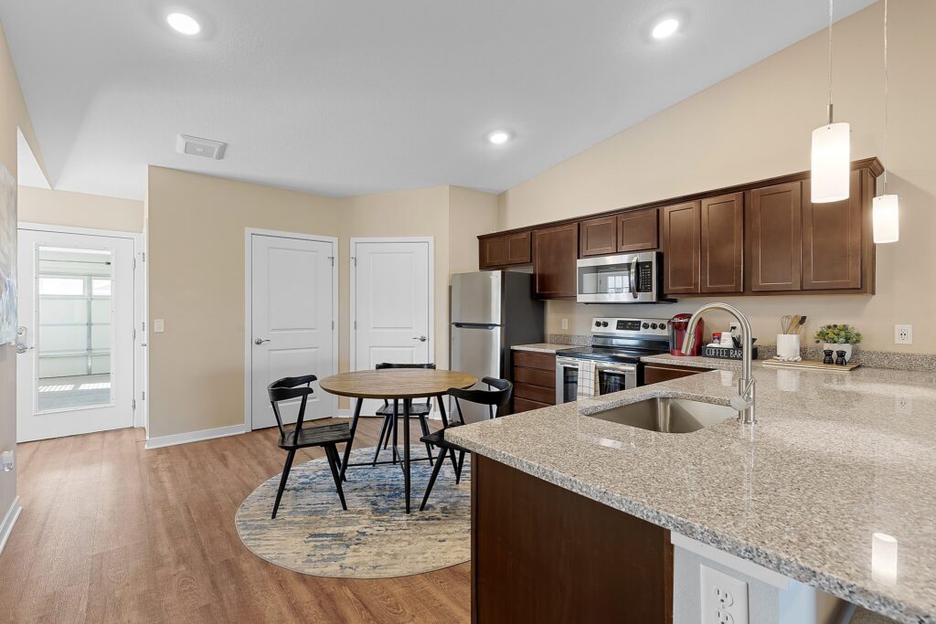 Modern kitchen and dining area with stainless steel appliances, round table with four chairs, granite countertops, and wood flooring. Double doors and entryway visible in the background.