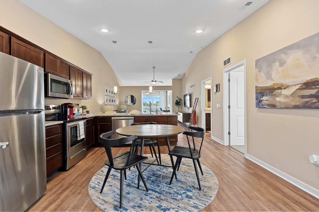 Open kitchen and dining area with stainless steel appliances, round table and four chairs, wood cabinets, hardwood floor, and wall art; living area visible in background.