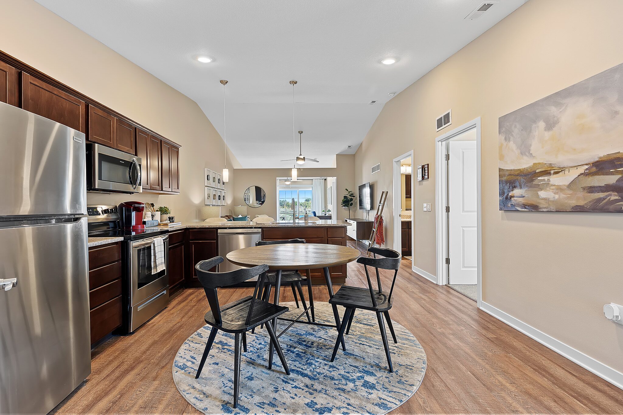 Open kitchen and dining area with stainless steel appliances, round table and four chairs, wood cabinets, hardwood floor, and wall art; living area visible in background.