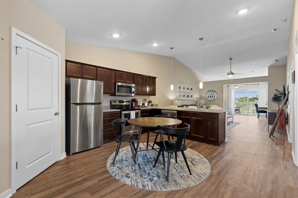 Modern kitchen and dining area with stainless steel appliances, dark wood cabinets, a round table with four chairs, and an open view to the living room and balcony.