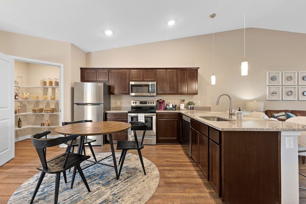 Modern kitchen with dark wood cabinets, stainless steel appliances, a round dining table with four chairs, and an adjacent open pantry stocked with jars.
