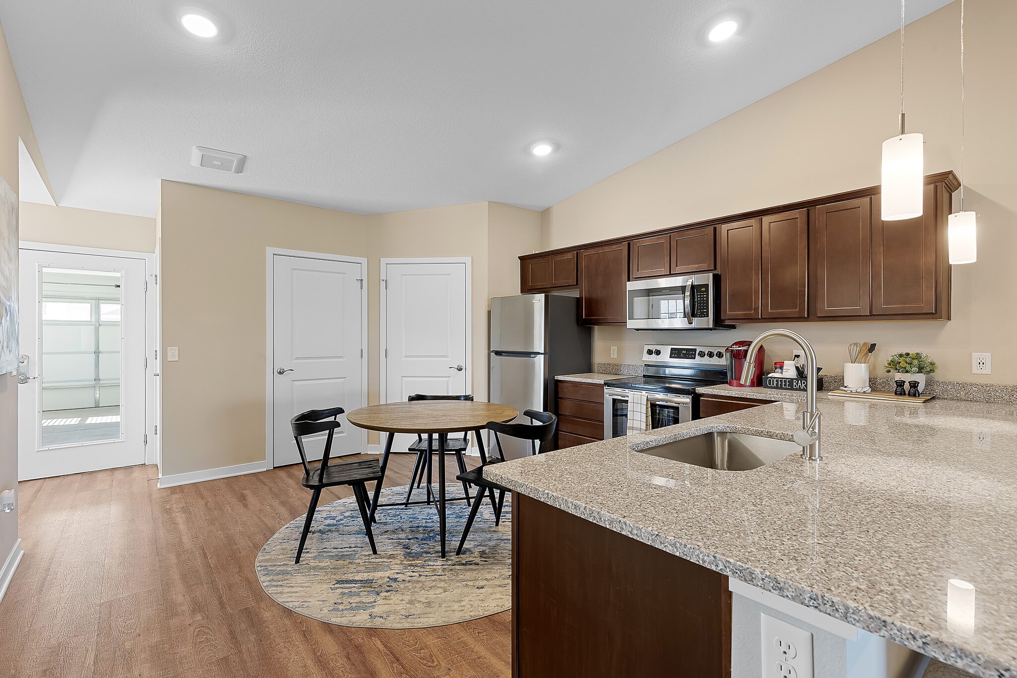 Modern kitchen and dining area with stainless steel appliances, round table with four chairs, granite countertops, and wood flooring. Double doors and entryway visible in the background.