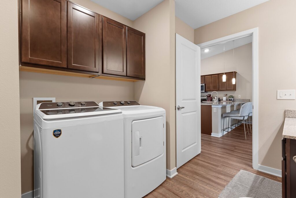 Laundry room with a white washer and dryer, brown cabinets above, beige walls, and a view into a kitchen with pendant lights and bar stools.