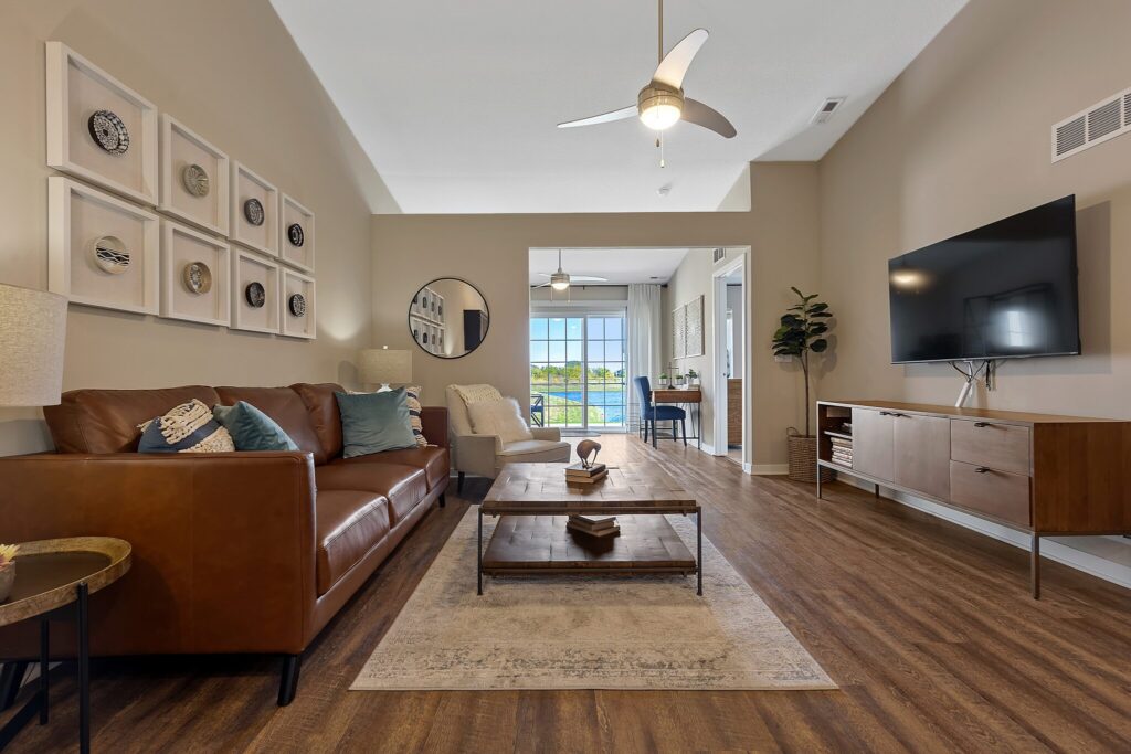 Modern living room with brown leather sofa, wooden coffee table, wall art, TV, and large window showing an outdoor view. Neutral colors and wood flooring create a cozy, contemporary space.