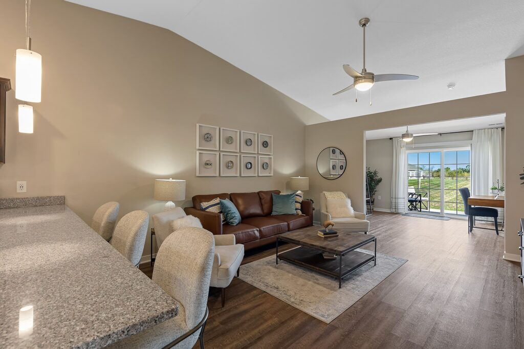 Open-plan living room with brown leather sofa, armchairs, coffee table, and dining area; large window with a view of greenery; neutral tones and modern decor.