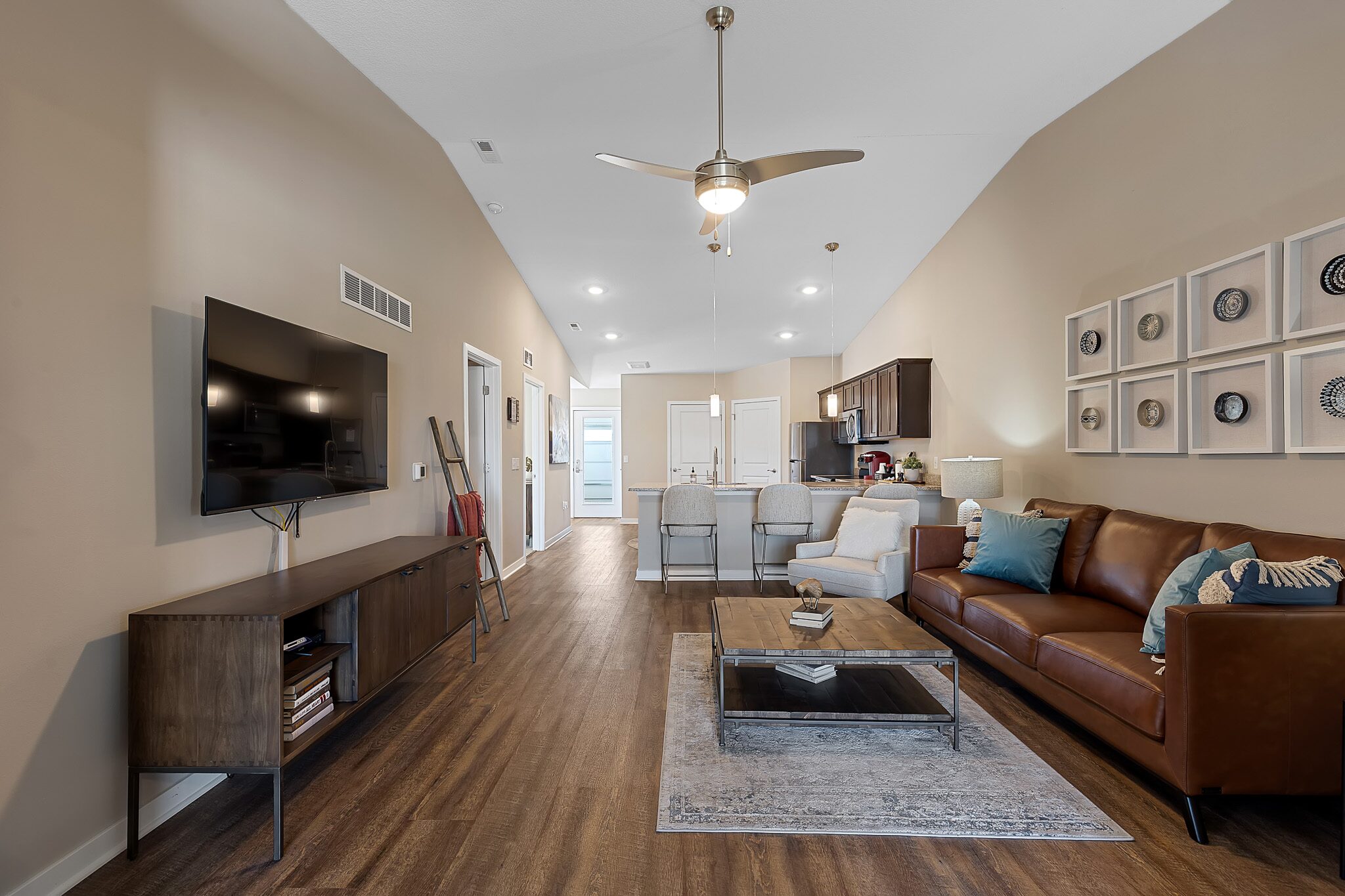 Modern living room with brown leather sofa, wall-mounted TV, wooden floors, open kitchen, and a ceiling fan, decorated with neutral tones and a gallery wall of framed plates.