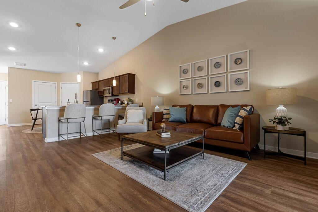 Modern open-concept living room and kitchen with brown sofa, armchair, bar stools, wooden floors, and neutral decor.