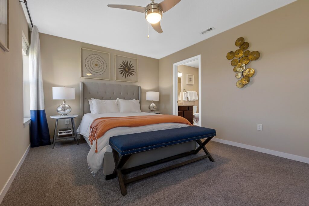 A neatly made bed with an orange throw blanket, two bedside tables with lamps, a bench at the foot, and a view into an adjoining bathroom.