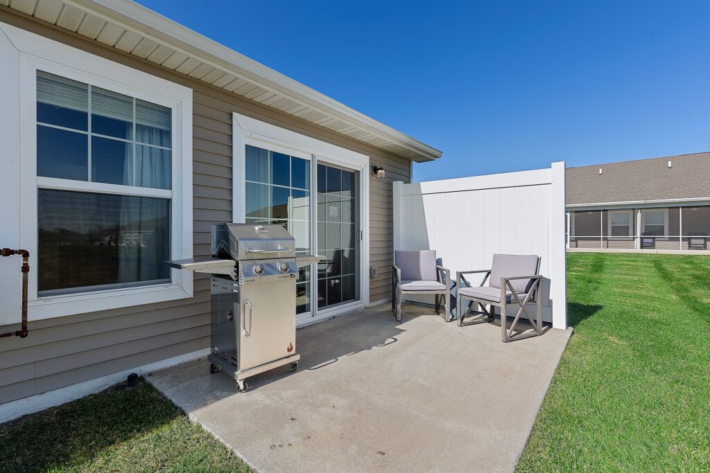 Concrete patio with two gray chairs, a small table, and a stainless steel gas grill beside a sliding glass door outside a beige house on a sunny day.