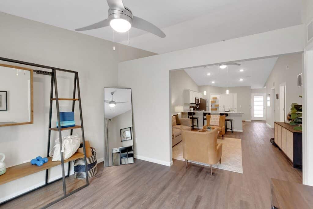 Modern open-concept living space with wood floors, neutral walls, ceiling fan, shelving, mirror, and view into kitchen and dining area with beige chairs and island.