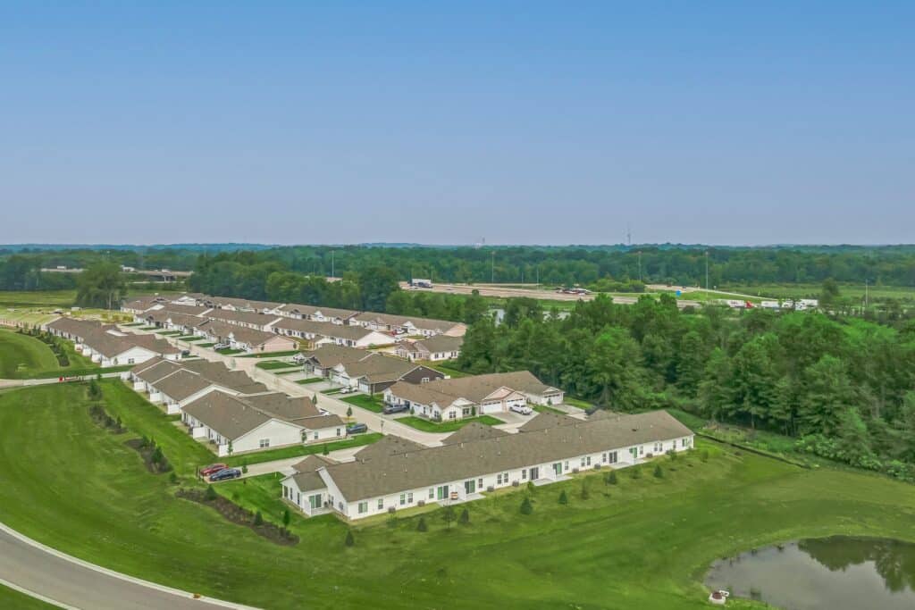 Aerial view of a suburban residential neighborhood with rows of single-story houses, green lawns, a small pond, and surrounding trees under a clear blue sky.