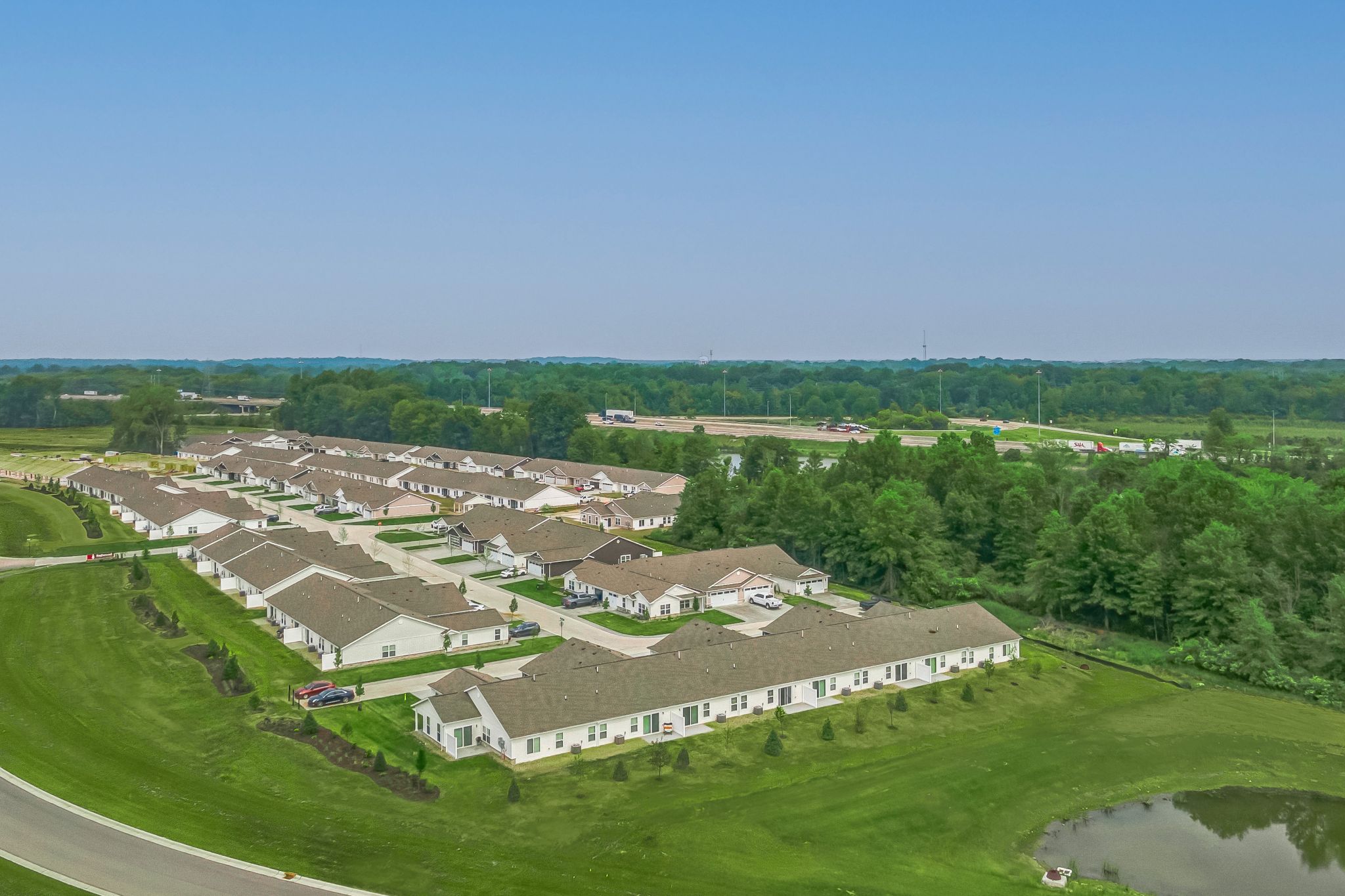 Aerial view of a suburban residential neighborhood with rows of single-story houses, green lawns, a small pond, and surrounding trees under a clear blue sky.