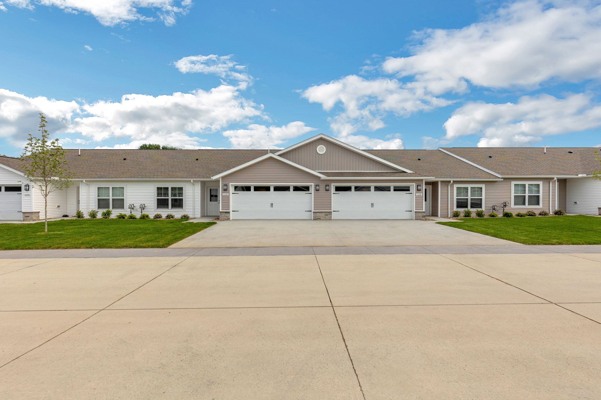 Single-story duplex homes with attached garages, white and beige siding, and neatly trimmed lawns under a partly cloudy sky.