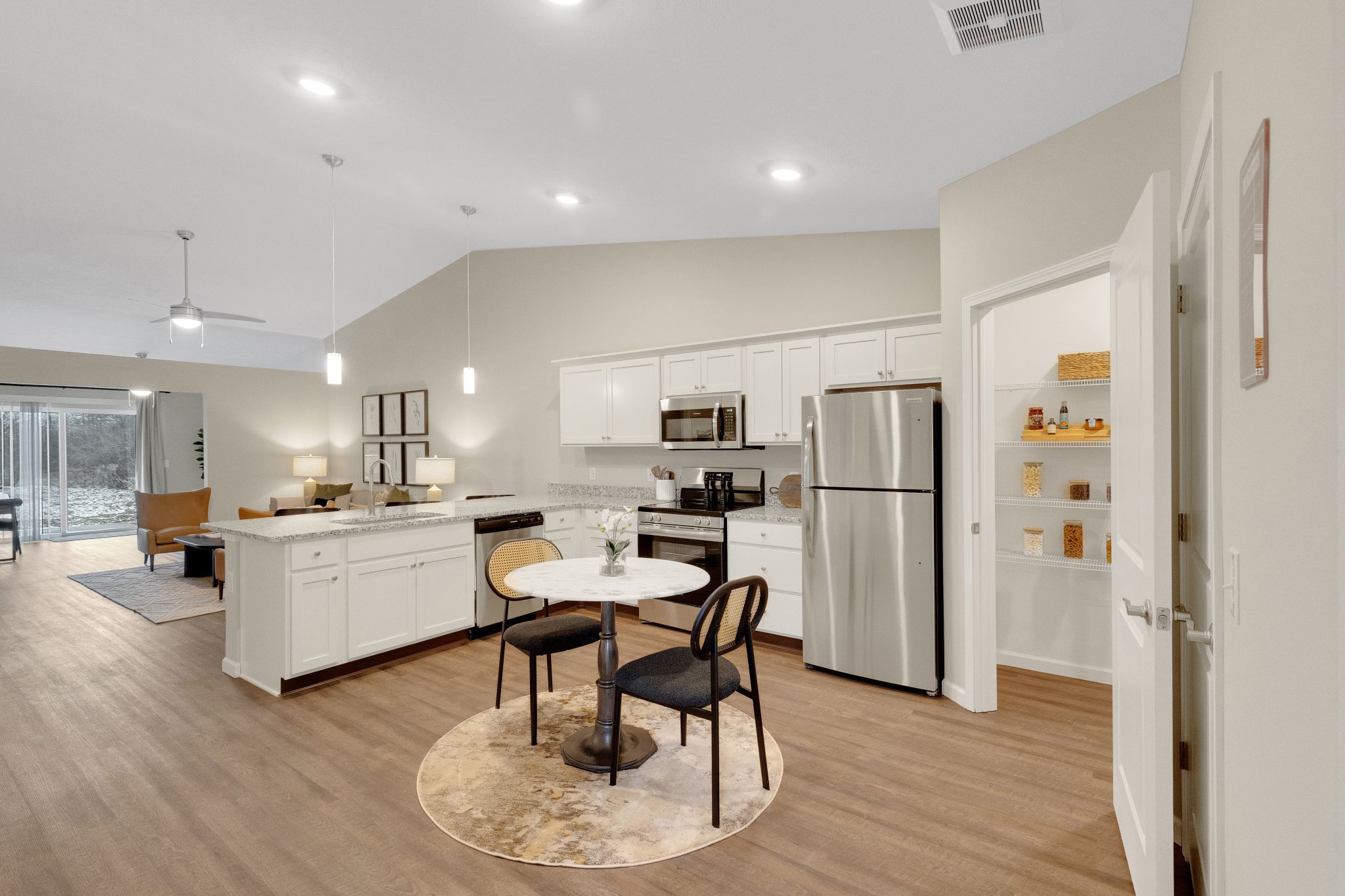 Modern kitchen with white cabinets, stainless steel appliances, a small round dining table with two chairs, and an open pantry stocked with jars.